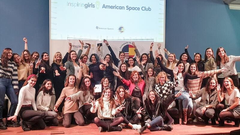 Se observa más de 20 chicas sentadas en el suelo con los brazos abiertos salundando a cámara. Detrás de ellas, una presentación en PowerPoint con las letras de Inspiring Girls.