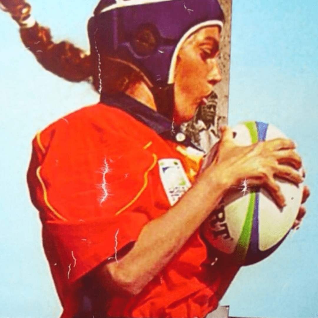 Se observa a una mujer de costado sujetando un balón de rugby. Viste una camiseta naranja d manga corta y un casco azul. Tiene el pelo recogido en una trenza castaña. El balón es ovalado, blanco, con dos rayas en el centro azul y verde.