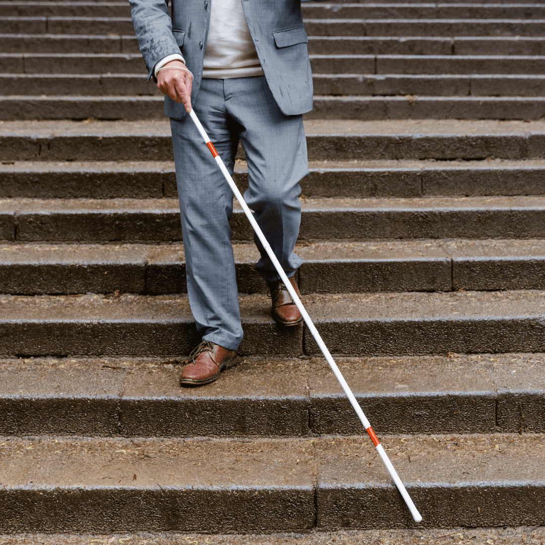 Se observan unas escaleras y la imagen de un hombre invidente caminando por ellas. Solo se ve desde el pecho hasta los pies. Lleva un traje gris y unos zapatos marrones. Tiene un bastón blanco con líneas rojas.