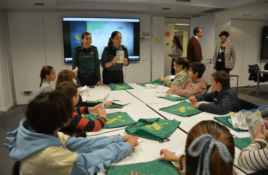Se observa dos mujeres castañas con el pelo recogido en coletas y uniformes de la guardia civil. Están enseñando un folleto a unos niños. Los niños tienen aproximadamente seis o siete años y están mirando atentos a las mujeres.