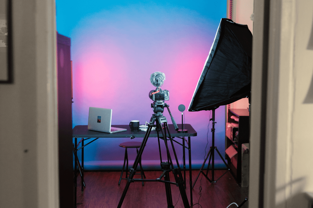 Estudio de fotografía Se observa un estudio de fotografía. Hay una pantalla verde al fondo. Delante tiene una mesa negra, con un ordenador blanco encima, un micrófono y una taza negra. Delante de la mesa hay una cámara negra con trípode. A la derecha de la fotografía hay un foco de luz negro grande.