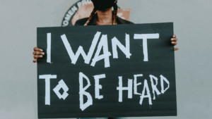 Se observa a una muchacha joven negra peinada con trenzas y con mascarilla negra sujetando un cartel de color negro. El cartel tiene escrito en letra blanco I want to be heard (quiero ser escuchada).