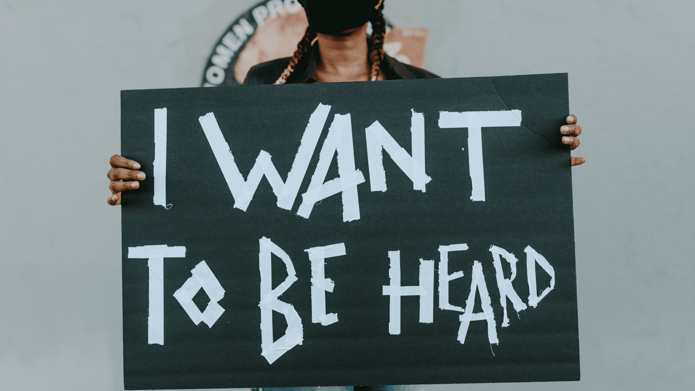 Se observa a una muchacha joven negra peinada con trenzas y con mascarilla negra sujetando un cartel de color negro. El cartel tiene escrito en letra blanco I want to be heard (quiero ser escuchada).
