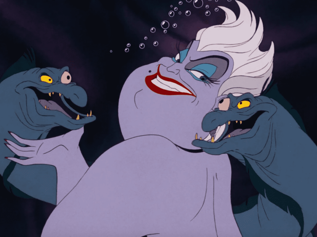 Imagen de la película animada 'La Sirenita' que muestra a Úrsula, la villana, con su piel púrpura, cabello blanco y labios rojos. Está sonriendo maliciosamente y sosteniendo a sus dos anguilas grises, Flotsam y Jetsam, una en cada mano.