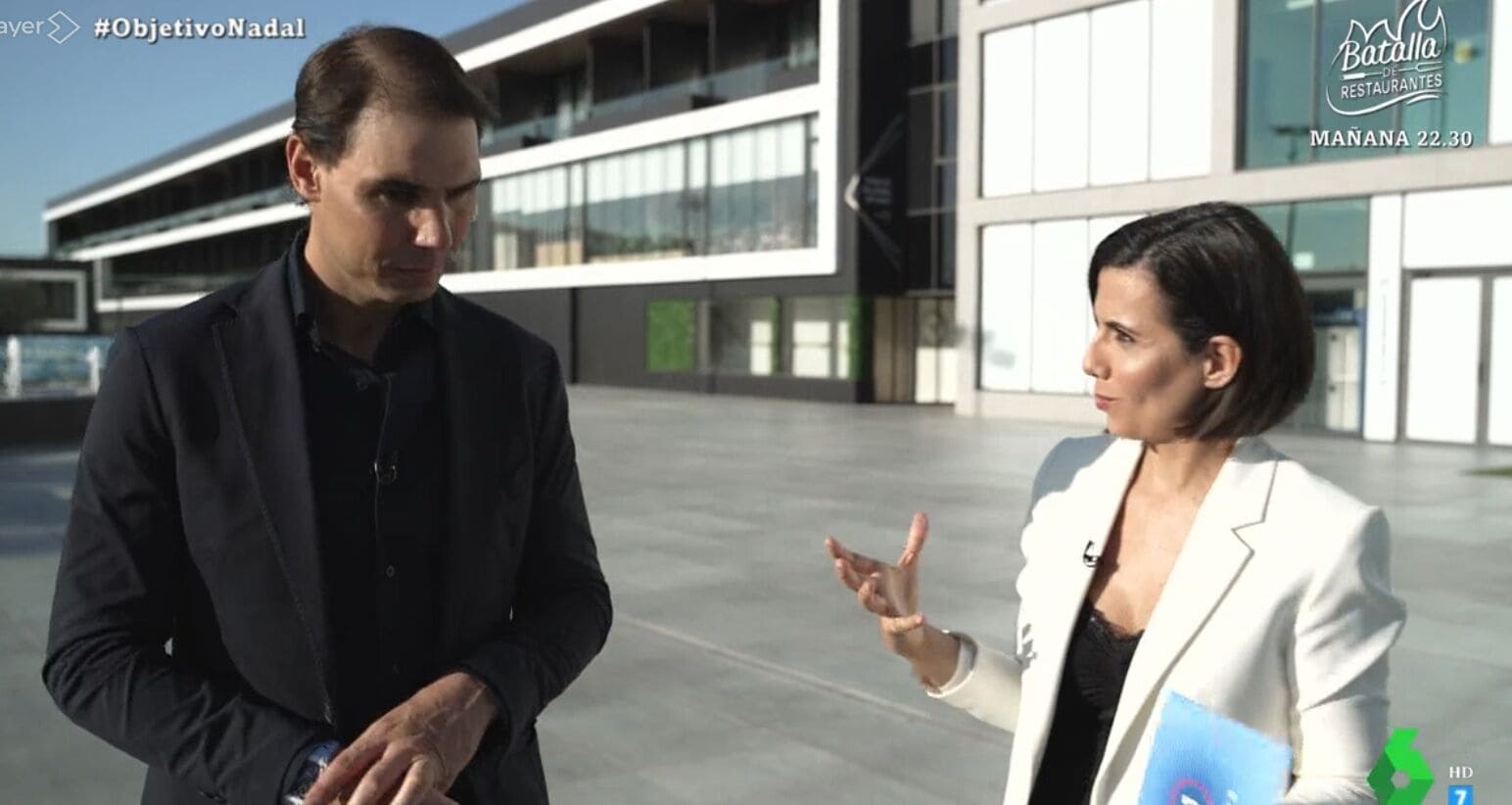 Se observa a Ana Pastor y Rafael Nadal conversando durante una entrevista en El Objetivo. Ana Pastor lleva el pelo negro corto, una chaqueta blanca y una blusa negra. Nadal lleva un traje engro.