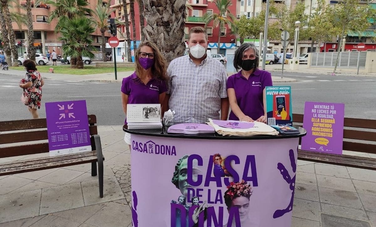 Se observa un stand redondo de color lila con el título en el centro, de color morado: Casa de la Dona. Encima del stand hay un calendario, un poster azul sobre la guía preventiva de violencia machista y varios abanicos de color lila. Detrás del stand, se sitúan dos mujeres con camisetas moradas y mascarillas. En medio de ellas, un hombre con camisa a cuadros y mascarillas.