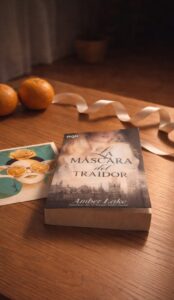 en un mesa de madera se observa el libro La máscara del traidor. A su izquierda una postal azul y naranja de Sevilla. Al fondo dos mandarinas